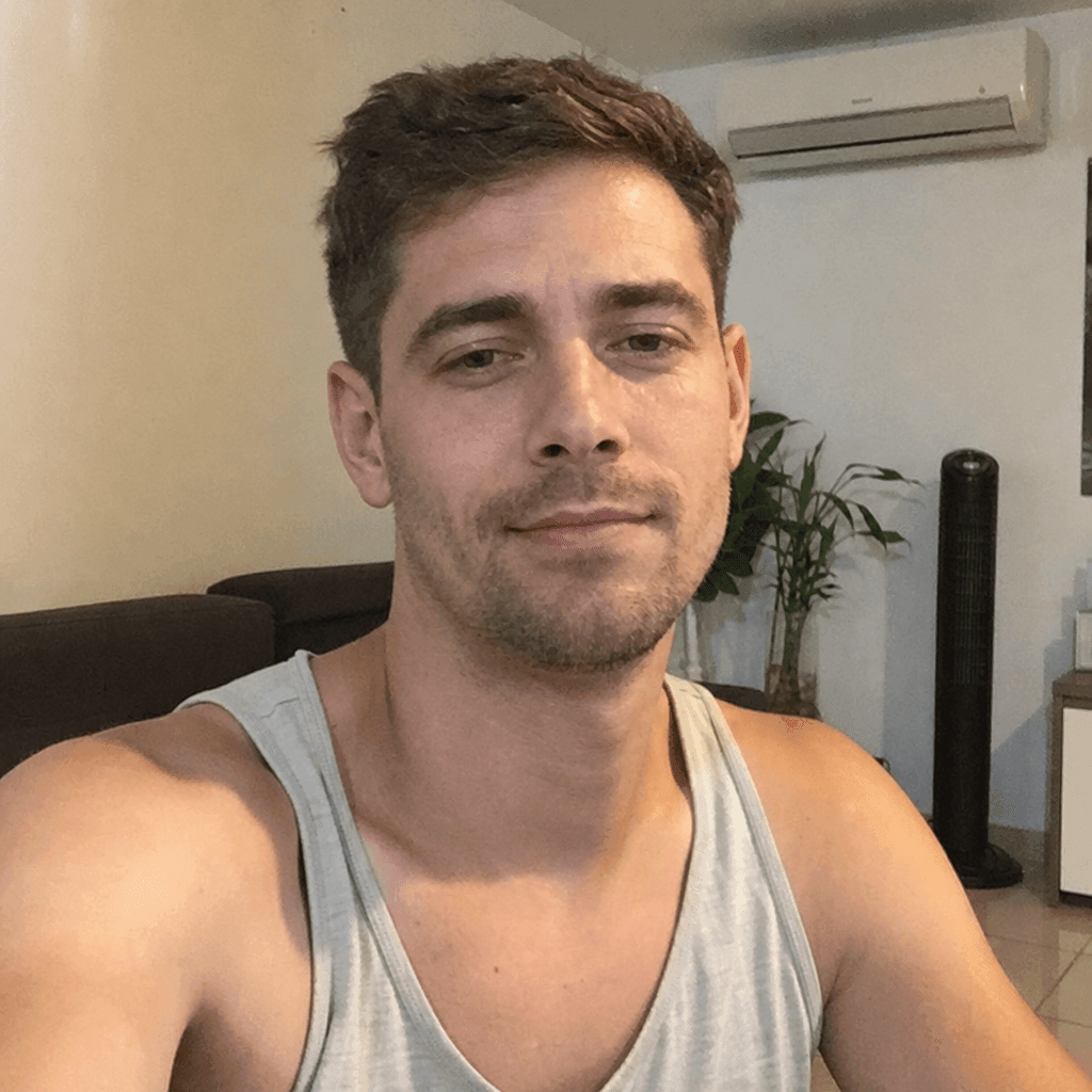 Homem branco jovem em regata cinza, selfie em sala brasileira com armário e papéis ao fundo — exemplo de foto informal